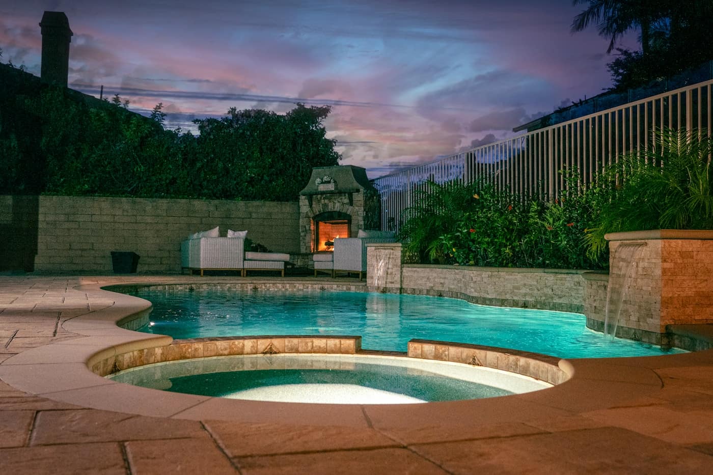 A backyard pool and spa lit at dusk beside a paver deck