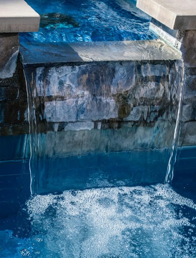 Raised spa spilling into a pool over a stone wall, with dark blue tile below the waterline